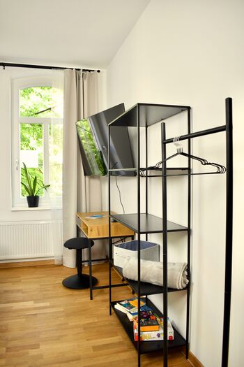 Zimmer 1: Zeigt den eingerichteten Arbeitsplatz mit einem darüber montierten Flachbildfernseher. Ein offenes Regal und eine Kleiderstange mit Bügeln bieten stilvolle und praktische Aufbewahrungsmöglichkeiten für Ihre Kleidung.