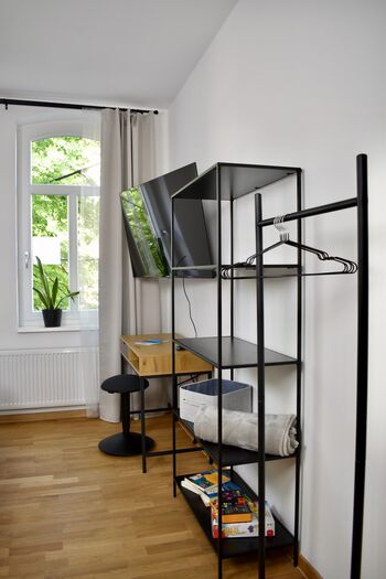 Zimmer 1: Zeigt den eingerichteten Arbeitsplatz mit einem darüber montierten Flachbildfernseher. Ein offenes Regal und eine Kleiderstange mit Bügeln bieten stilvolle und praktische Aufbewahrungsmöglichkeiten für Ihre Kleidung.