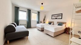 Modernes Doppelzimmer mit zwei Einzelbetten, Schlafsofa und stilvoller Einrichtung. Helle Atmosphäre durch große Fenster, gemütliche Beleuchtung und dekorative Akzente – ideal für entspannte Aufenthalte.