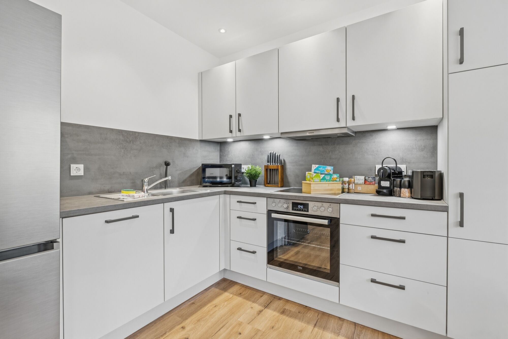 Weiße L-Küche mit grauer Arbeitsplatte, Backofen, Mikrowelle, Fenster mit Grünblick.