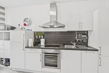 Weiße Küche mit dunkler Rückwand, Ceranfeld, Backofen, Wasserkocher und Wanduhr.