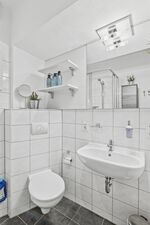 Weiß gefliestes Badezimmer mit Duschkabine, Waschtisch, Schminkspiegel und Pflanzen auf Wandregal.