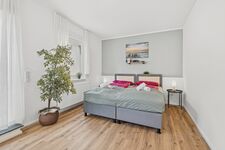 Doppelbett mit grauer Bettwäsche, große Zimmerpflanze, heller Holzboden, schlichte Wanddeko.