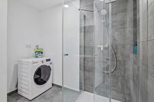 Graue Fliesendusche, Einbauwaschmaschine daneben, moderne Armaturen, helle Beleuchtung.